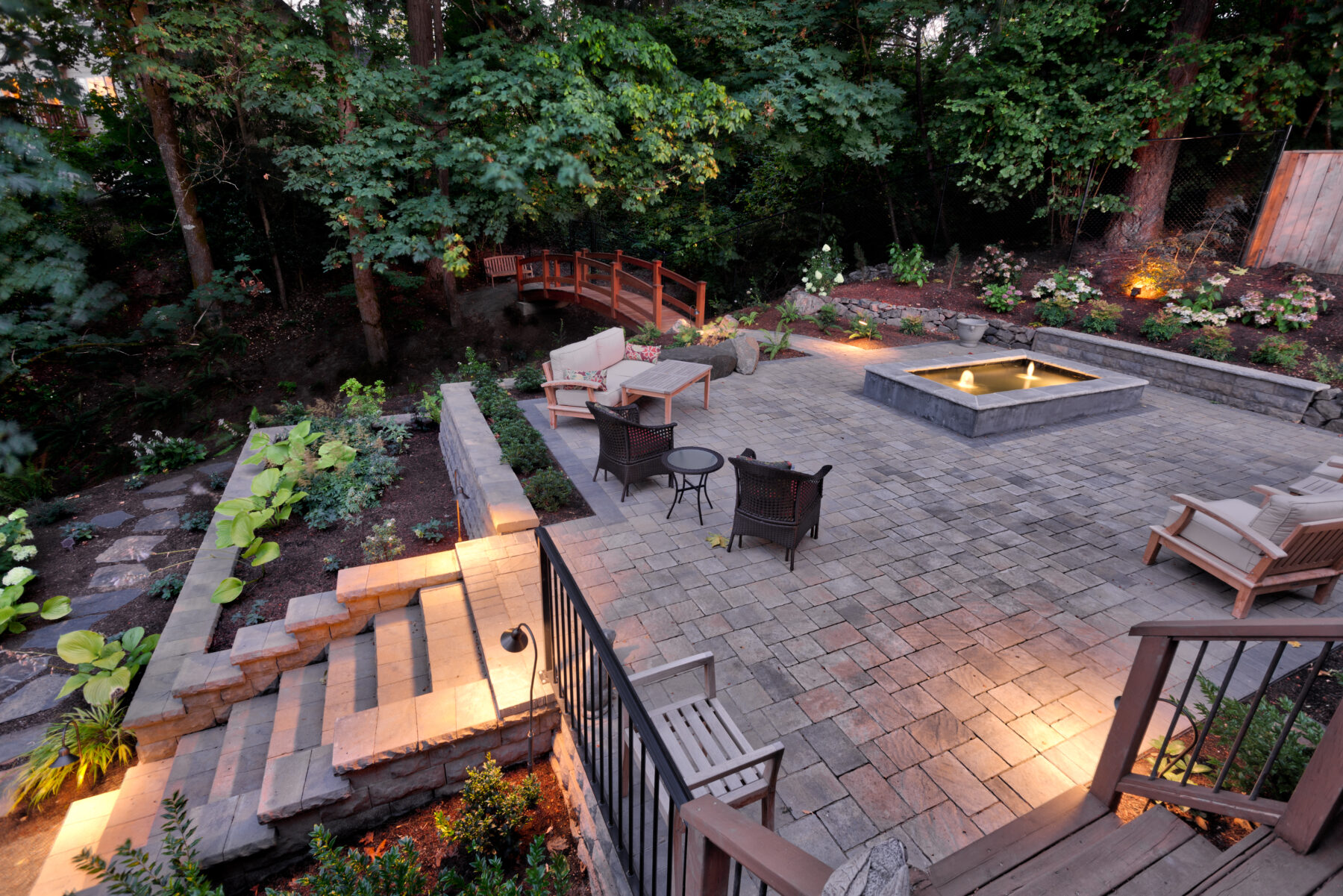 Illuminated evening hardscape patio with fire pit and outdoor furniture set amidst a lush Pacific Northwest garden, ideal for hosting and social gatherings