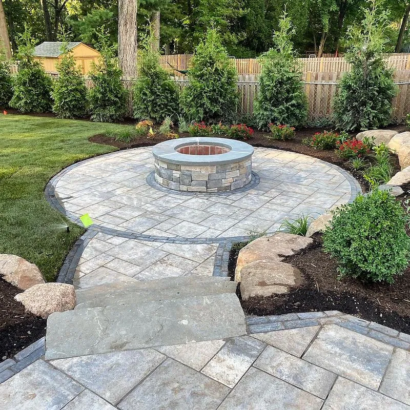 Elegant circular paver patio design with central fire pit, bordered by lush greenery and privacy fencing, embodying outdoor tranquility for entertainment and relaxation